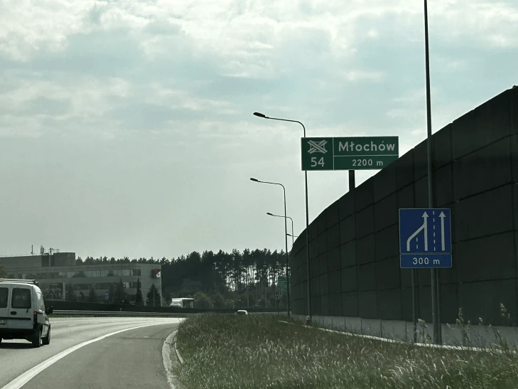 Zjazd z drogi szybkiego ruchu z widocznym znakiem kierunkowym „Młochów 2200 m” oraz barierami akustycznymi po prawej stronie.