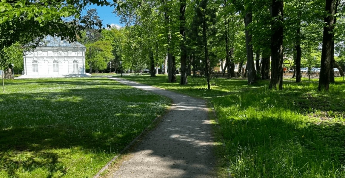 Park z alejką spacerową otoczoną drzewami, w tle widoczny zabytkowy budynek.